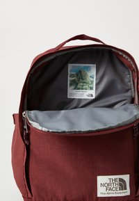 Burgunderfarget ryggsekk med grå innside, som har en liten bildeetikett av Yosemite inni. Glidelåsåpning og slitesterk tekstur.