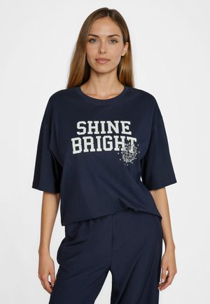 Woman with long brown hair wears navy blue oversized shirt with "SHINE BRIGHT" text and decorative beads, paired with matching navy pants.