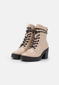 Bottines en cuir verni beige avec lacets noirs, talon épais et semelle en caoutchouc robuste. Présente des détails de couture et des accents en métal.