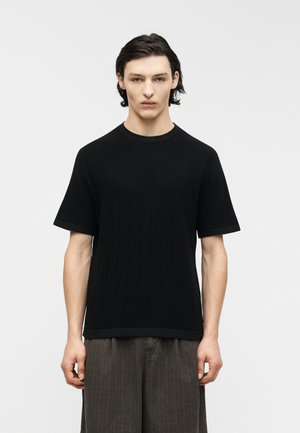 Giovane uomo con capelli neri corti, che indossa una maglietta nera a maniche corte lavorata a coste e pantaloni largi grigio scuro, in piedi davanti a uno sfondo bianco.