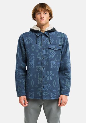 Junger Mann trägt eine blaue, gemusterte Jacke mit Knöpfen, mit schwarzer Kapuze und weißem Fleecefutter, steht vor einem einfarbigen Hintergrund.