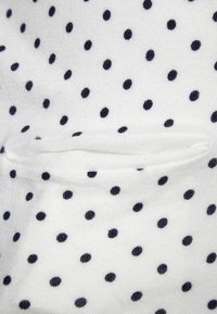 Vitt tyg med ett upprepat mönster av små marinblå prickar, med en diskret slits eller fickdetalj synlig i texturen.