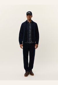 Marineblaue Cordjacke über einem karierten Hemd, kombiniert mit passenden marineblauen Hosen und braunen Schnürschuhen, abgerundet mit einer schwarzen Kappe.