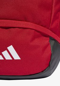 Crveni ruksak od tkanine s crnom donjom stranom, s istaknutim bijelim logom s tri pruge. Materijal izgleda izdržljivo s teksturiranom površinom.