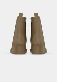 Um par de botas de tornozelo em camurça beige com saltos empilhados e alças traseiras, mostradas de costas sobre um fundo branco.
