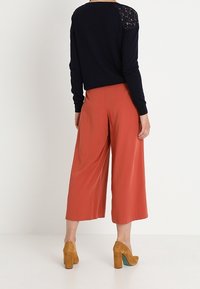 Pull marin avec des détails en dentelle sur les épaules, associé à un pantalon large couleur rouille et des chaussures à talons blocs beiges. Texture lisse et coupe décontractée.