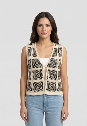 Femme portant un gilet en tricot sans manches beige et noir à motifs, noué à l'avant, par-dessus un haut blanc et un jean bleu clair, devant un fond uni.