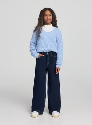 Ragazza con capelli ricci che indossa un maglione azzurro peloso, dolcevita bianco, jeans larghi scuri e sneaker bianche, in piedi con una mano in tasca.