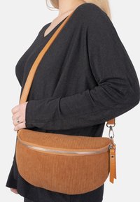 Braune Corduroy-Crossbody-Tasche mit strukturiertem Oberflächen, silbernem Reißverschluss-Detail und einem taubenledernen Trageriemen, getragen über einem schwarzen Oberteil.