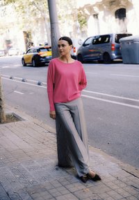 Suéter rosa de manga larga de punto con escote redondo, combinado con pantalones de pierna ancha de color gris claro y zapatos marrones de punta afilada, con un fondo de ciudad.