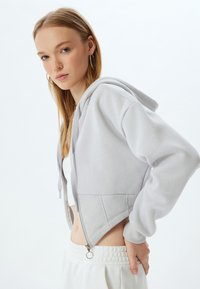 Hoodie grigio corto con dettaglio zip all'orlo, realizzato in un morbido tessuto testurizzato e dotato di cappuccio con cordino. Abbinato a pantaloni bianchi.