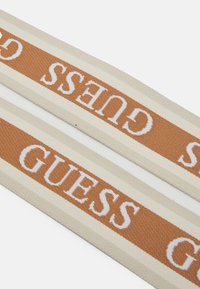 Tkany materiałowy pasek o beżowej bazie z brązowymi akcentami i białym logo "GUESS". Gładka faktura z poziomymi paskami.