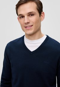 Marineblauer V-Ausschnitt-Pullover aus Strickmaterial, mit geripptem Kragen und Bündchen, getragen über einem weißen T-Shirt mit einem dezenten Markenzeichen.