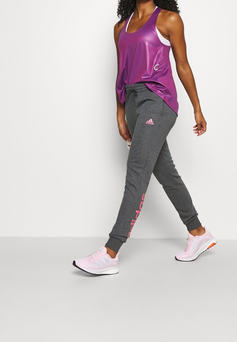 Paarse mouwloze sporttop, grijze joggers met roze accenten en lichtroze hardloopschoenen. Kenmerken zijn een elastische tailleband en logo-details.