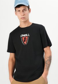 Sort bomulds t-shirt med korte ærmer, prydet med et rødt emblem med et løvedesign og teksten "O'Neill" i hvidt. Cremefarvede detaljer på logoet.