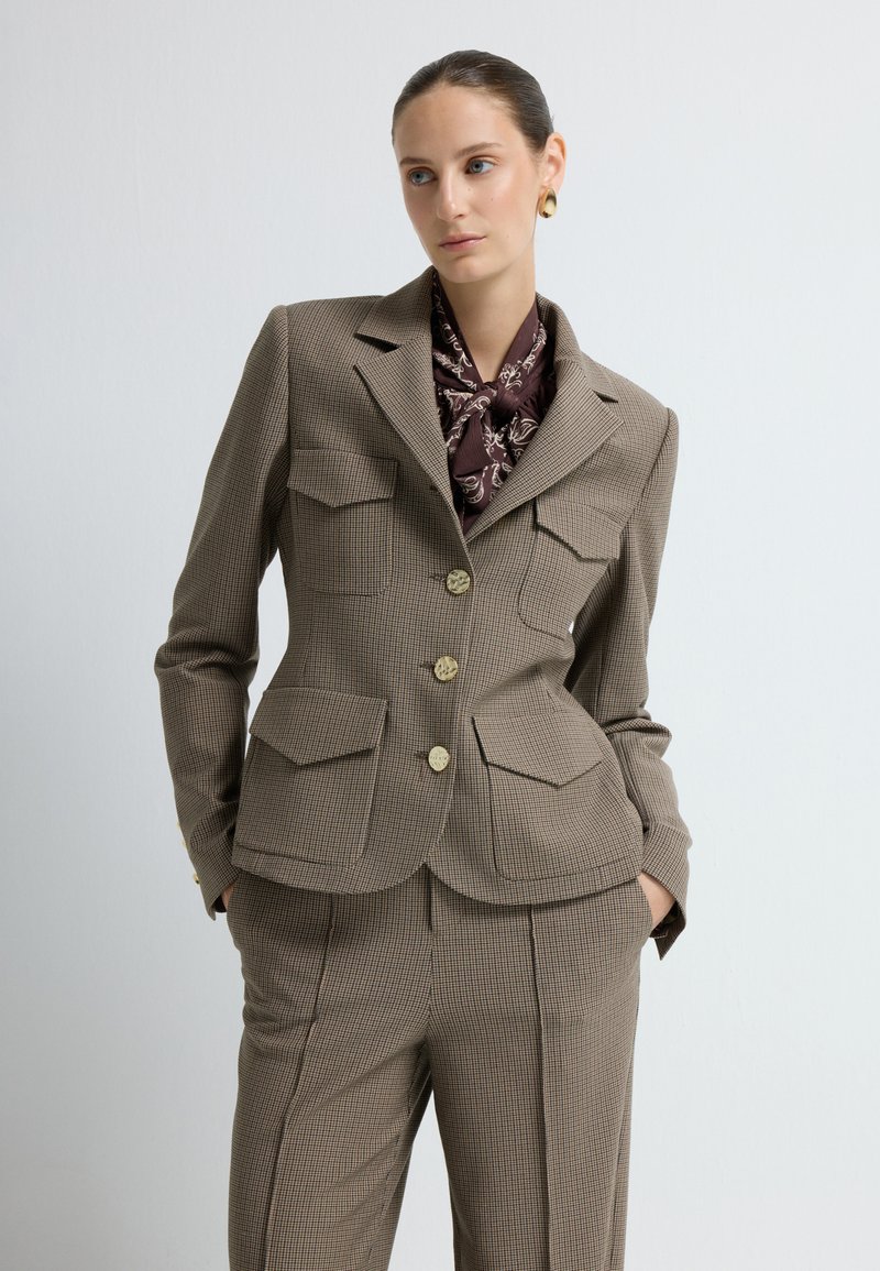 Brun ruderblazer med to forlommer, guldknapper og en notch-lapel. Båret med et mønstret brunt tørklæde for at tilføje ekstra detalje.