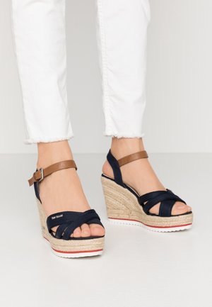 Fødder iført marineblå og brune kile-sandaler med flettede platformsåler og hvide bukser med frynset kant.