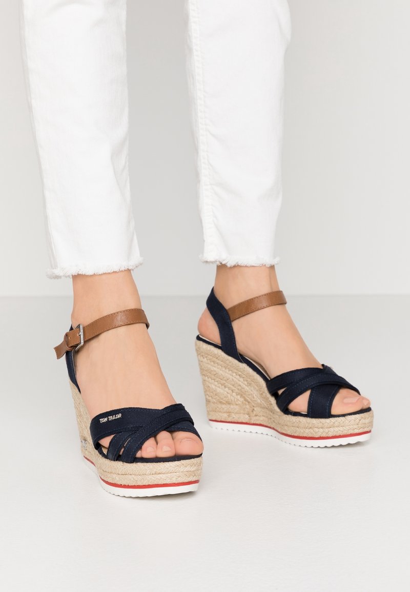 Pieds portant des sandales compensées bleu marine et marron avec semelles tressées et un pantalon blanc avec des ourlets effilochés.