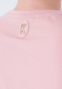 Camiseta de algodón rosa con escote redondo y un parche ovalado texturizado con un logotipo estilizado de "K" en la parte trasera del cuello. Acabado suave.