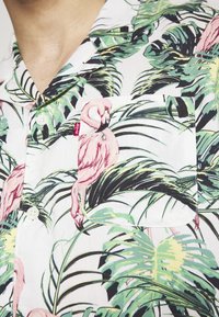 Camicia a maniche corte con bottoni, tessuto bianco con motivi di foglie tropicali verdi e fenicotteri rosa. Presenta una tasca e un'etichetta di marca.