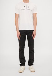 Man in een wit Armani Exchange T-shirt, zwarte broek en witte sneakers, staand tegen een effen lichte achtergrond.