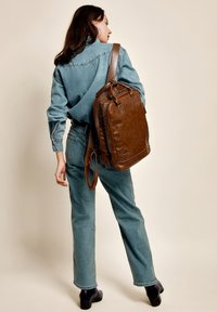 Smeđi kožni ruksak s teksturiranim završetkom, s patentnim zatvaračem i dvostrukim ručkama; naočit s plavom denim košuljom i trapericama.