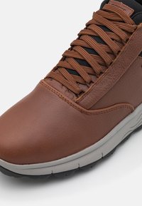 Brązowe skórzane sneakersy z teksturowanym wykończeniem, z ciemnobrązowymi sznurowadłami i szarym gumowym spodem. Wysoka cholewka z wzmocnionymi szwami.