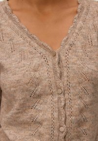 Cardigan en tricot beige clair avec un col festonné, des motifs de feuilles détaillés et quatre boutons ronds en argent sur le devant.