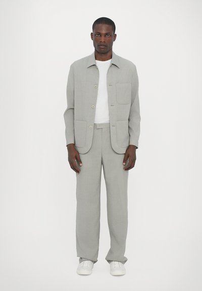 Γκρίζο, ελαφρύ κοστούμι από ύφασμα με σακάκι που κουμπώνει, περιλαμβάνοντας δύο μπροστινές τσέπες και αντίστοιχες ευθείες παντελόνες, συνδυασμένο με λευκά αθλητικά παπούτσια.