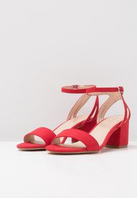 Sandales à talon bloc en daim rouge avec un design à bout ouvert, une bride de cheville et un intérieur lisse. Présente des accents minimalistes pour un look épuré.