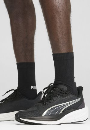 Schwarze Sportschuhe mit Mesh-Obermaterial, silbernen Akzenten und weißer Sohle. Kombiniert mit schwarzen gerippten Socken, die am Bund ein Logo tragen.