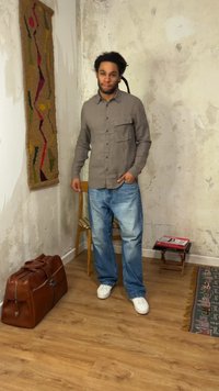 Mann mit Dreadlocks steht in einem Raum, trägt ein graues Hemd mit Knöpfen, blaue Jeans und weiße Sneakers in der Nähe von einer Ledertasche, einem Holzstuhl und einem kleinen Tisch.