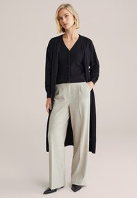 Cardigan lungo nero sopra un top a coste nero con scollo a V, abbinato a pantaloni grigio chiaro a gamba larga e tacchi neri a punta. Texture lisce, vestibilità moderna.