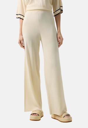 Kobieta ubrana w kremowe spodnie o szerokich nogawkach oraz sandały na platformie z odkrytymi palcami, stojąca na tle jednolitego tła.