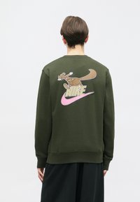 Sweatshirt verde com decote redondo e punhos canelados. Apresenta um canguru em cartoon e o logótipo da Nike em rosa e dourado nas costas.