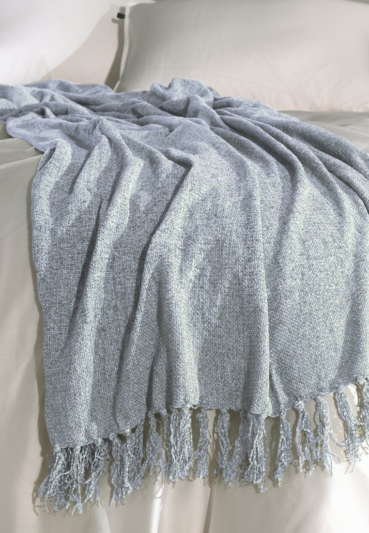 Texturovaná, světle modrá deka přehozená přes postel, se třásněmi na okrajích a tkaným vzorem, umístěná na krémovém povlečení.