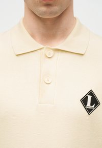 Gros plan d'une personne portant un polo crème avec deux boutons fermés et un écusson noir en forme de losange avec le logo "Lacoste" sur la poitrine.