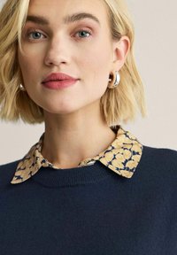 Mörkblå stickad tröja med rund hals, lager-på-lager över en gul blommig skjorta med krage. Modellen har kort blont hår och stora silverfärgade hängande örhängen.