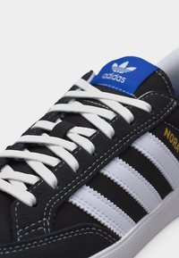 Μαύρο αθλητικό παπούτσι Adidas με λευκά κορδόνια, τρεις λευκές ρίγες στο πλάι, μπλε ετικέτα λογότυπου στη γλώσσα και "NORA" με χρυσά γράμματα.