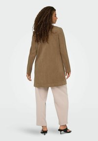 Abrigo largo de ante marrón con mangas largas, combinado con pantalones beige claros y sandalias de tacón negras. Textura suave y diseño de corte relajado.