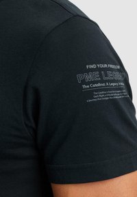 T-shirt nera in cotone con maniche corte. Stampa sulla spalla sinistra che include elementi testuali e grafiche di colore bianco a contrasto.