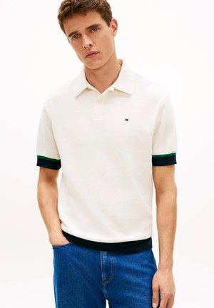 Giovane uomo che indossa una polo bianca a maniche corte con rifiniture verdi e nere sulle maniche e sull'orlo, abbinata a jeans blu, in piedi con una mano in tasca.