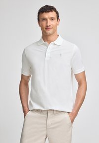 Camisa polo blanca con un cuello clásico, mangas cortas y un pequeño logo en el lado izquierdo. Llevada con pantalones beige claros.