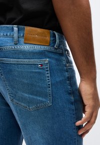 Δεξιά πλευρά ατόμου που φοράει μπλε τζιν Tommy Hilfiger με ορατή πίσω τσέπη, ετικέτα με το λογότυπο της μάρκας και καφέ δερμάτινη ετικέτα στη ζώνη μέσης.