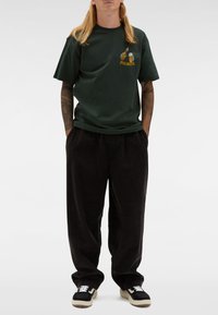 Osoba z długimi blond włosami, ubrana w ciemnozieloną koszulkę z grafika piwa, luźne czarne spodnie oraz czarno-białe trampki, z rękami w kieszeniach.