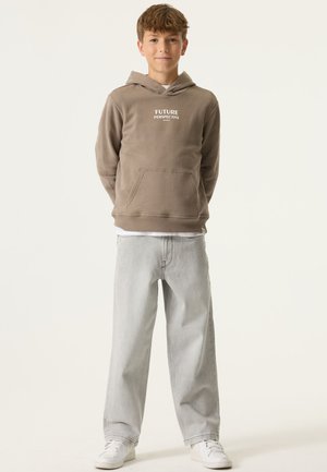 Bruine hoodie met de tekst "FUTURE PERSPECTIVE", voorzak; gecombineerd met lichtgrijze wijdvallende jeans en witte sneakers. Effen achtergrond.