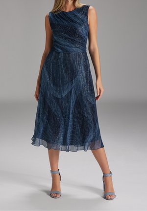 Kobieta w granatowej sukience midi bez rękawów z abstrakcyjnym połyskującym wzorem i wysokich sandałach na obcasie z paskiem wokół kostki, stojąca na jasnoszarej podłodze.