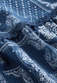 Niebieska tkanina z białymi wzorami paisley i geometrycznymi, z ozdobnym tkanym koronkowym wykończeniem na marszczonym szwie.