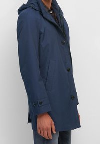 Námořnická modrá pláštěnka s kapucí, vyrobená z hladkého materiálu, s dvěma předními kapsami a černými knoflíky na manžetách a předním zapínání.