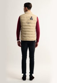 Chaleco acolchado beige con diseño de rombos, que presenta tres parches bordados en la parte posterior, usado sobre una camiseta de manga larga burdeos y jeans oscuros.